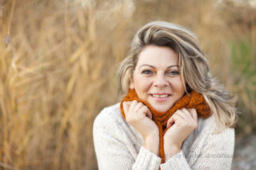 Eine mittelalte Frau mit blonden Haaren hält einen beigen Schal um ihren Hals und schaut dabei lächelnd in die Kamera.