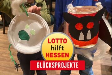 Zwei Kleinkinder halten selbst gebastelte Laternen hoch. Darüber das Logo von LOTTO hilft Hessen - Glücksprojekte.
